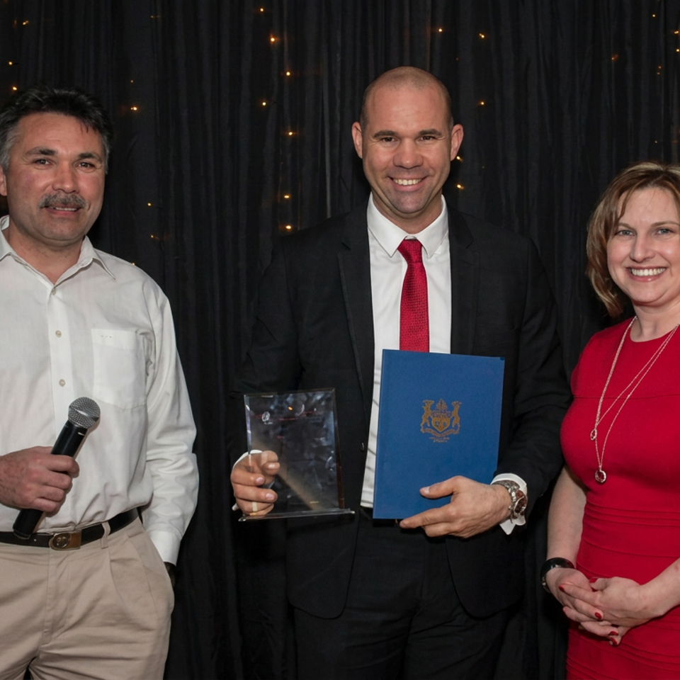 Three people pose at an awards event, with a man holding a microphone and another holding a trophy and blue certificate.